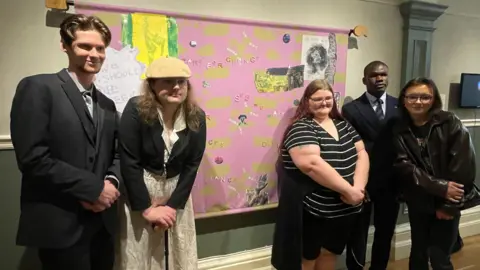Five young people standing in front of a banner full of phrases and questions about identity. The background is purple and green with slogans written across it.