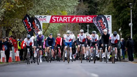 Paul Campbell A large group of riders head off from the start line of the Etape Loch Ness in Inverness. There is a large banner with the words "start" and "finish" in white letters on a red background.