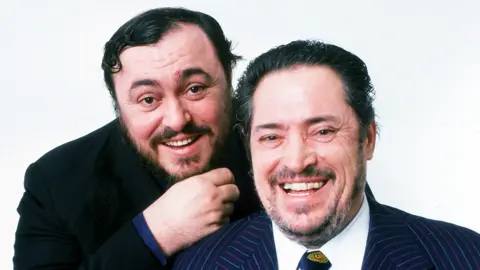 Getty Images Head and shoulders shot of Luciano and Fernando Pavarotti. Luciano is to the left and slightly behind his father. He has dark hair and a beard, and wears a black jacket with a navy blue shirt sleeve visible at the cuff. Fernando has dark slicked back hair with a few grey strands and a greying beard. He wears a white shirt and navy tie with a crest at the knot, and a navy suit with red and white thin pinstripes. Both men are smiling at the camera.