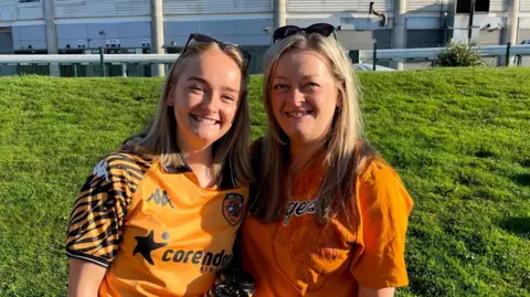 Richard Madden/BBC A mother and daughter, both with shoulder-length, blonde hair, are smiling. They are both wearing black and amber coloured Hull City shirts. Behind them is a football stadium. 