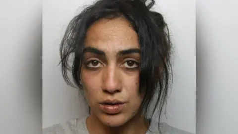A mugshot of a woman with long black hair, partly tied back, and wearing a grey top.