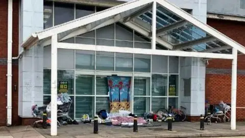 A white metal and glass canopy over the front of what looks to be a 1980s retail unit. It is shuttered behind the glass frontage, with a collection of bedding on the floor and personal possessions in shopping trolleys.