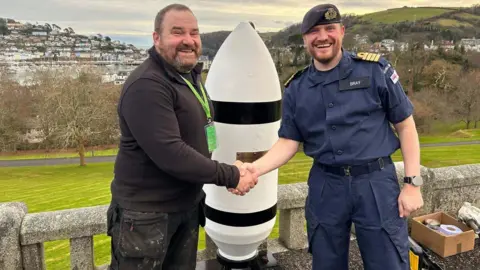 Dressed in black, master wheelwright Greg Rowland shakes the hand of Captain Andy Bray, who is dressed in a uniform of a dark blue short-sleeved shirt and trousers, plus a naval beret with badge on it.
They are standing in front of large white bomb shell, which is half the height of Mr Roland. It has a plaque on its casing. The town of Dartmouth is in the background.