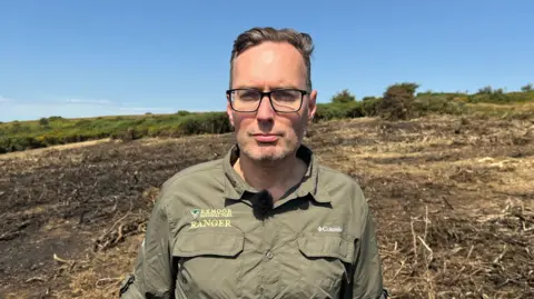 Tim Parish is wearing a khaki park ranger's shirt. He is wearing glasses and is looking into the camera. Behind him is a burnt field with green shrubbery in the distance.