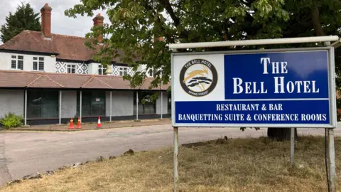 A white building is stood on the left with an entrance to the right which says The Bell Hotel, Best Western. A tree is in front of the two-storey building with three traffic cones outside