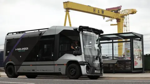 PA Media A small silver bus that has the branding 'Harlander' on the side. The yellow H&W cranes can be seen in the background.