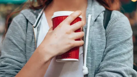 Getty Images Wanita mengenakan tudung abu-abu memegang cangkir kopi merah sekali pakai di dekat dadanya