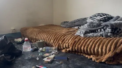John Fairhall/BBC A mattress on the floor of a messy room above a shop. It has a brown and grey sheet on top of it. Next to it are discarded water bottles, cigarettes and tissues.