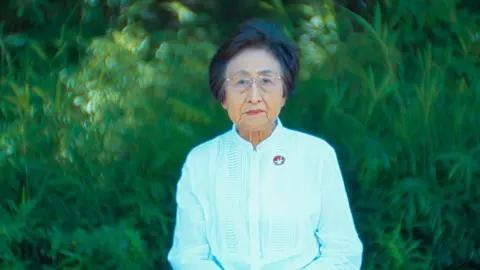 BBC/Minnow Films Portrait of Michiko Kodama, an elderly woman with short dark hair. She is wearing metal-rimmed glasses and has a serious expression. She is pictured standing in front of a green bush.