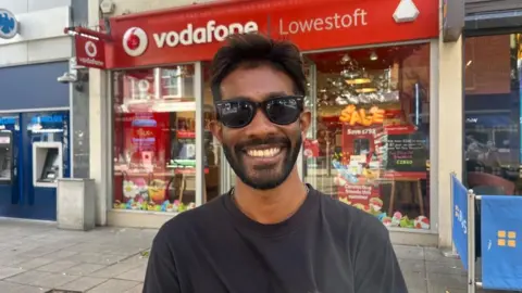 Andrew Turner/BBC Jeevan Hewa Mainaththuge is wearing sunglasses and a long-sleeved black T-shirt. He is smiling, standing outside the Vodafone shop in Lowestoft.