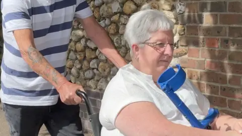 A woman, Pamela Bent, who has short grey hear, is wearing a white top and sitting in a wheelchair. She is holding a walking stick with a blue handle. A person wearing a blue striped top and grey jeans is pushing her in the wheelchair.