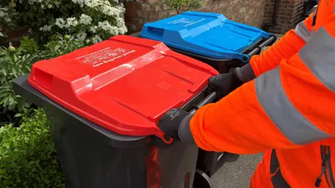 A worker in an orange hi vis jacket approaches two household recycling bins, one red and one blue.
