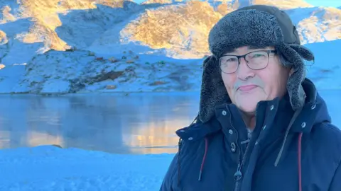 Kaaleeraq Ringsted in a black jacket and hat smiling in front of the fish with the fjord in the background.