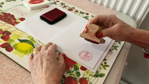 A woman's hands, painted with red nail varnish, holding a stamp. She has just pressed it on to a white envelope, leaving a round red post mark reading "Lover".