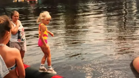 Meredith Ingbretson A blond child stands on a log in the water, balancing in socks, sneakers, pink shorts and a pink and yellow top, her blonde hair tied up in a ponytail