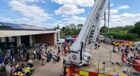Bedfordshire Fire & Rescue Service A large amount of people at a fire station open day, complete with a white crane extended. There are vehicles about and a large building to the left. 