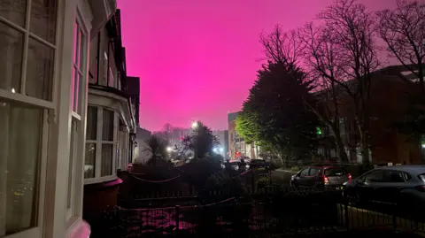 Gareth Glover Pink sky over a street with terraced houses on the left and parked cars and trees on the right.