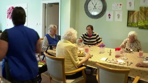 The image shows a group of older people sitting around a large dining table, eating a meal together in a bright communal room with light green walls, framed artwork and a wall clock. A care worker is visible moving around the table.