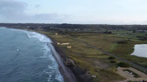 BBC Aerial view of a coastline with a sea wall, coastal road, wetland lagoon and nearby buildings in St Ouen.
