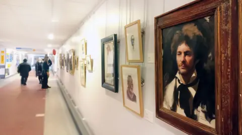 A gallery wall with various artwork in different coloured frames. The one in the foreground is a painting of a man with brown messy hair and a black suit and black tie around his white shirt. There are people further down the corridor looking at the artwork.