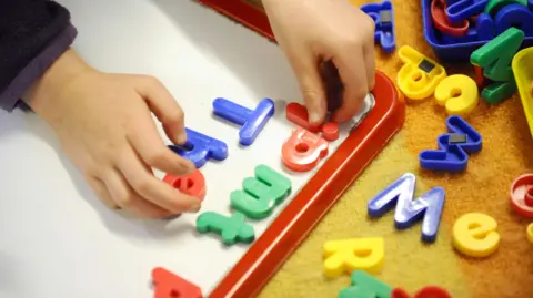 A child plays with lettered fridge magnets. The letters are blue, green, yellow and red. The child is arranging them on a white board. 