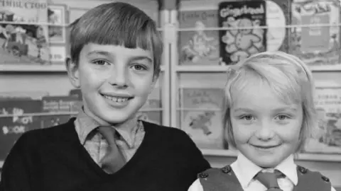 Mayall Family A black-and-white photo of a boy with brown hair, brushed forward over his forehead. He is wearing a shirt and tie under a dark v-neck jumper. He is standing next to a younger girl with blonde hair, wearing a shirt and tie under a sleeveless top. They are standing in front of shelves displaying children's books.
