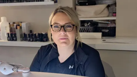 A woman with blonde hair sits behind a manicure station in a salon, wearing a black zip‑up top. Shelves behind her hold bottles of nail polish, beauty products, and neatly stacked supplies. A desk lamp and manicure tools are visible in the foreground.
