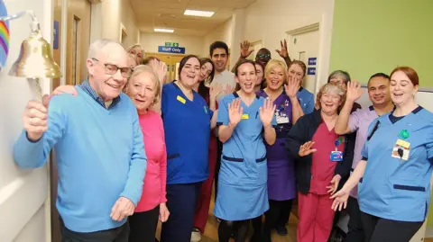 Kettering General Hospital A large group of people in a hospital corridor, smiling and waving at a camera. Bill Clarke, to the left is holding a bell, with his wife, Alison next to him. She is looking at him and Bill is looking at the camera and smiling. 