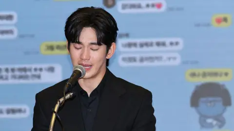 A tearful Kim Soo-hyun looking down and speaking into a microphone. He is wearing a black shirt and black blazer. Behind him is a projection of screenshots of text messages.