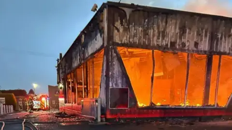 Orange flames engulf a building. The windows of the building have been smashed and black suit covers the outside walls. On the left of the image a fire truck and crew can be seen. There is a large hose on the ground. 