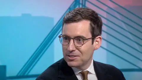 Dan Price, wearing a black suit, white shirt and brown tie, talks in the BBC's Salford studio.