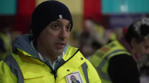 David Goodfellow wearing a high-visibility jacket and a black beanie speaks inside a busy hall, with other volunteers in similar clothing working in the background.