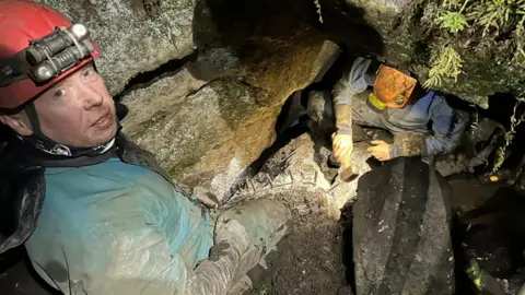 Irish Cave Rescue Organisation A cave rescuer with a red helmet and blue clothes stands inside a cave. Below him is a man in a yellow helmet and gloves, he is holding a hammer and banging against the floor of the cave.