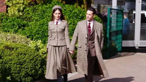 PA Media A young woman wears a tweed hat, jacket and full-length tweed skirt, white shirt and red tie. She holds the hand of a man in a matching tweed overcoat, tweed jacket, red tie and white shirt. 