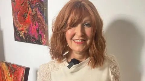 Elaine Murray stood next to her paintings, one of them behind her is red and pink and blue and brown.