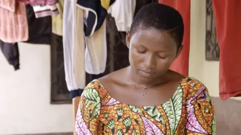 BBC A woman with short hair and a patterned dress glances down.