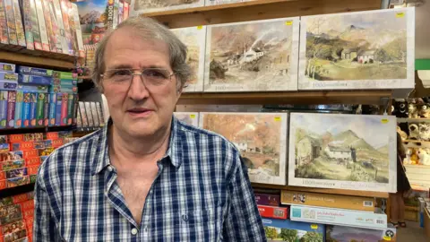 John Lowther standing on from of lots of jigsaw puzzles, many showing countryside paintings with mountains and trees in them. He has grey hair and is
wearing a blue and white checked shirt.