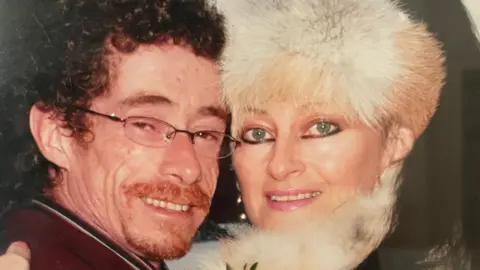 Stephen Alexander Man and woman hugging while smiling at the camera. Male has dark curly hair and a ginger beard wearing glasses. Female has white fluffy hat and scarf on.