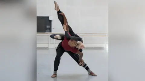 Andre Uspenski Melissa dancing with with José Alves in a dance rehearsal studio. She is wearing a black unitard and white ballet shoes. José is wearing a red t-shirt and black trousers. He is bent forward holding Melissa on his back. She is grasping his right arm and left leg, he is grasping her left leg and right hand.