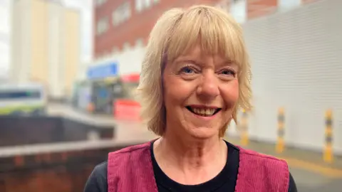 BBC / Elizabeth Baines A woman with blonde hair smiles at the camera. She wears a black top with red straps. Behind her the St James' emergency department is blurred.