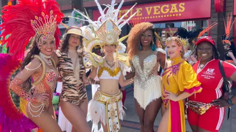 Lily-May Symonds/BBC Women wearing different carnival outfits stand together to pose for a picture. Some of them wear large headpieces complete with feathers and other decorations that tower above them.