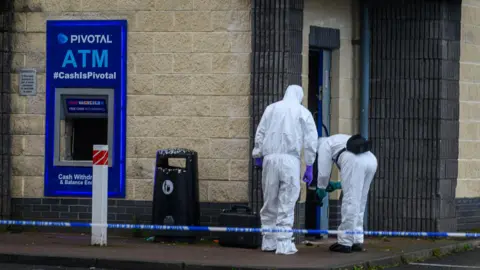ATM missing from wall of shop with men in white suits investigating. 