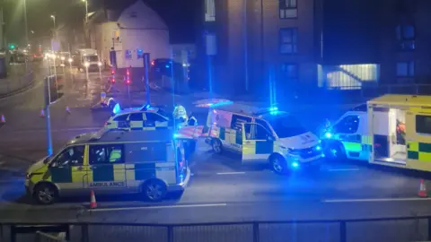 Three ambulance and a police car with their emergency lights on at the scene of a crash at a junction in a town centre. Part of the road has been cordoned off by cones. Buildings surround the junction.