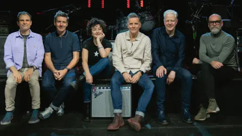 Jamie Carter Deacon Blue, a band, sitting in a row. There is an amplifier in the centre of the group and instruments behind them. 