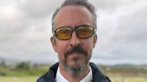 Paul Logue has grey hair and a grey beard. He is looking into the camera. He has dark rimmed glasses and is wearing a white shirt, dark spotty tie and dark jacket.
