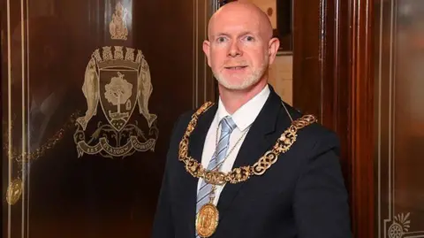 A bald man wearing a large gold chain, black suit jacket, white shirt and blue striped tie stands next to a wooden door which has the gold-embossed Glasgow coat of arms on it