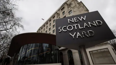 The outside of New Scotland Yard. The name of the station is written in silver letters on a black board outside a large cream building