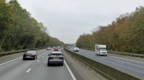 View of a dual carriageway from the traffic going southbound. There are mulitple vehicles driving across the two lanes either side of the carriageway. Rows of trees line up beside the motorway.