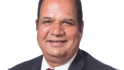 Councillor Chaman Lal in an official photo. He is wearing a suite, smiling and there is a white background behind him.