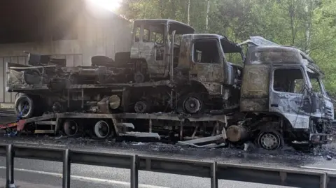 Charred shell of lorry with a further two being carried on its trailer.  The lorry is at the side of a road under a bridge